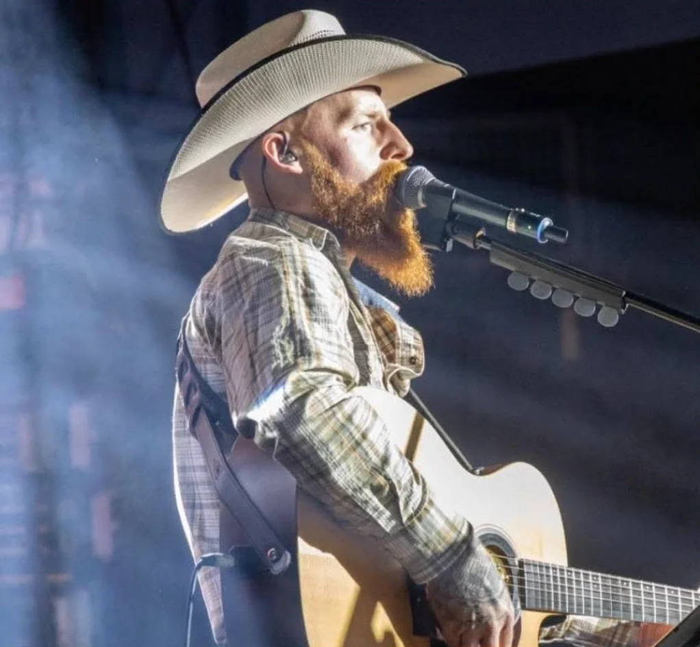 A man with a red beard wearing a cowboy hat, plaid shirt, playing an acoustic guitar and singing into a microphone on stage.