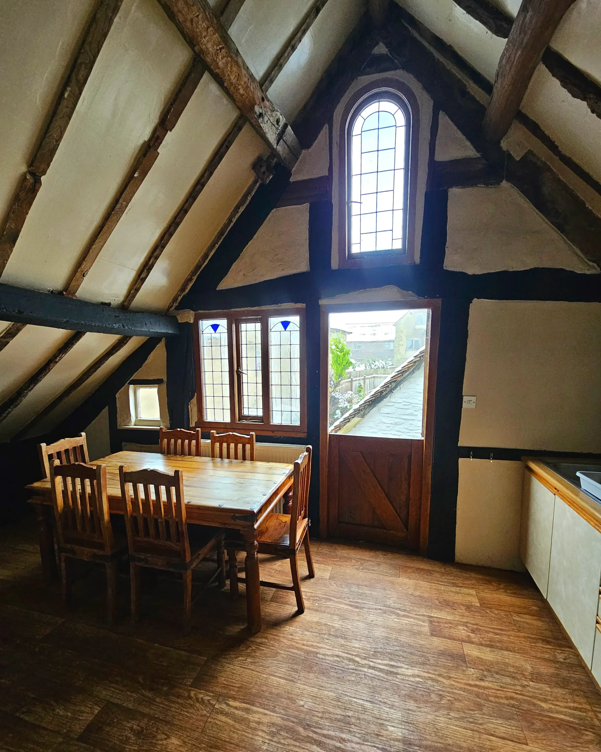 An indoor room with wooden beams on the ceiling, a large window with stained glass, a wooden dining table with chairs, and a view of the opposite house and sky outside.