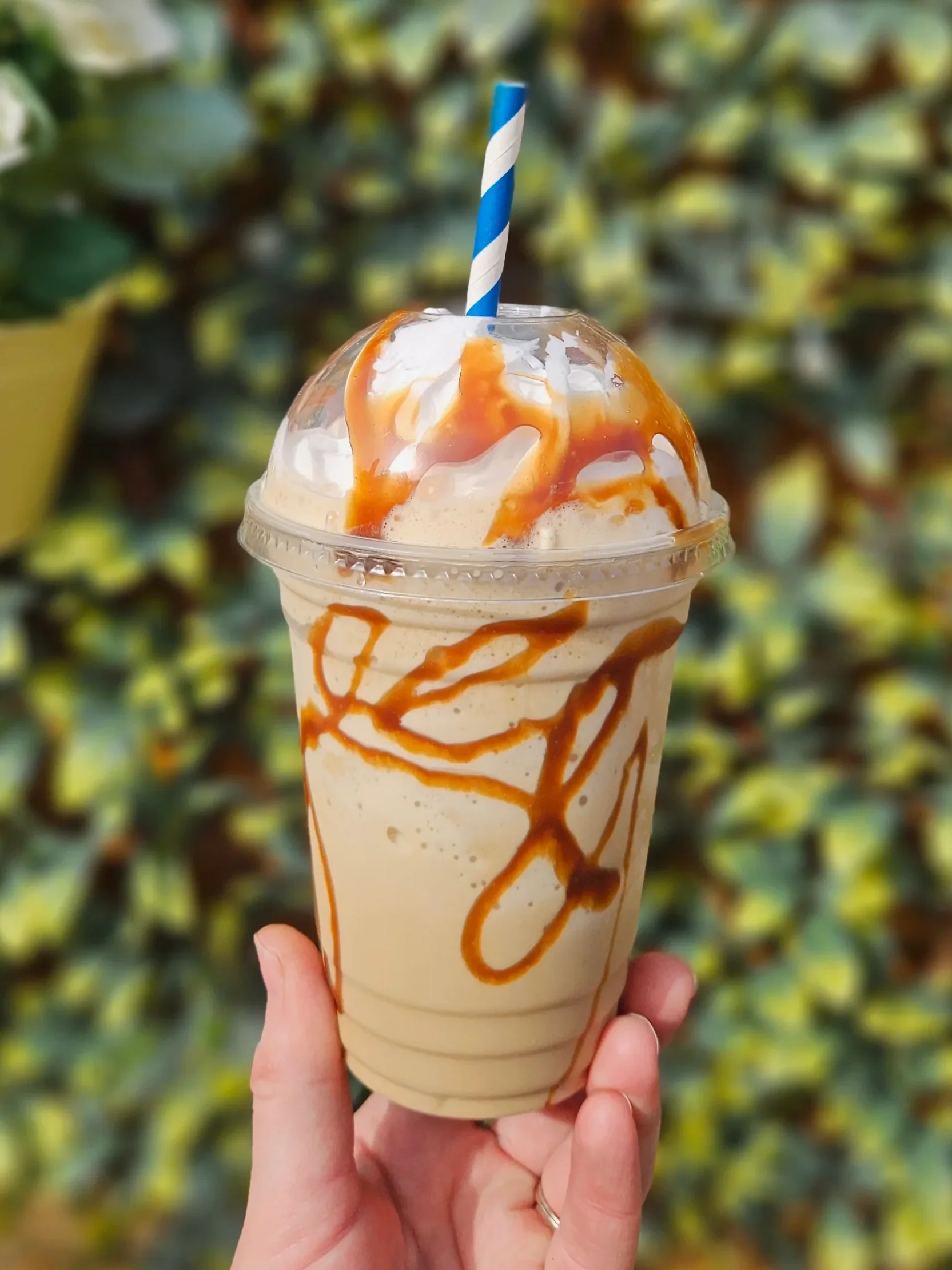 A person holding a milkshake with caramel syrup drizzle and a blue and white striped straw in front of a blurred background of yellow and green plants.