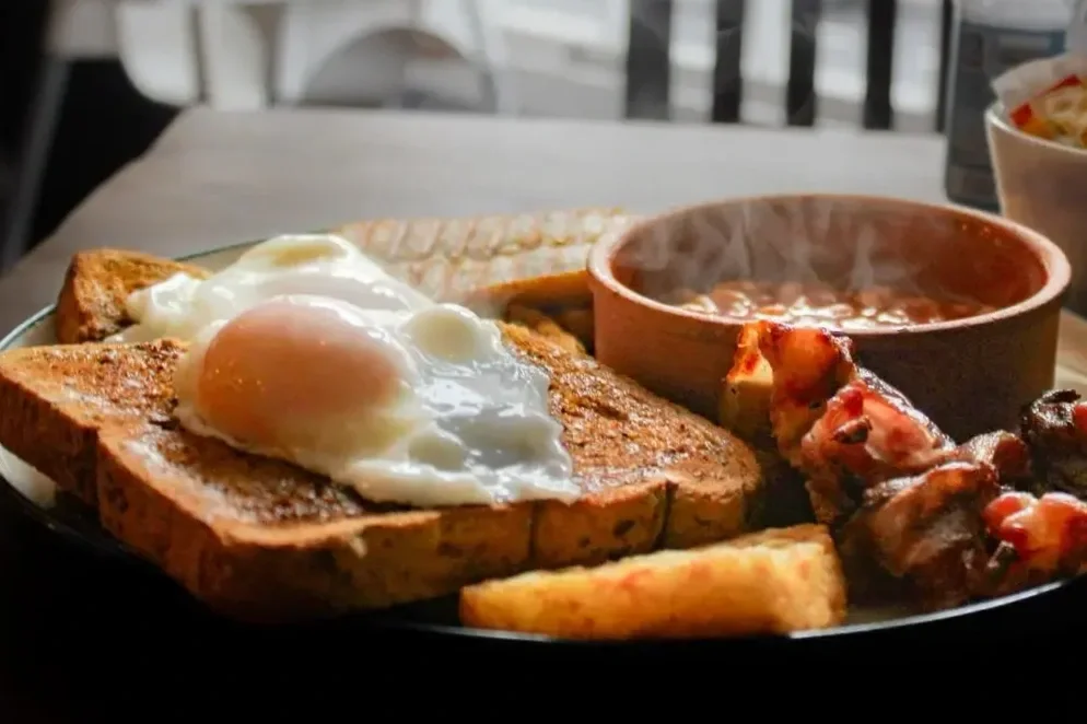 A breakfast plate with toast topped with a fried egg, a bowl of baked beans, grilled chicken wings, and a piece of fried plantain.