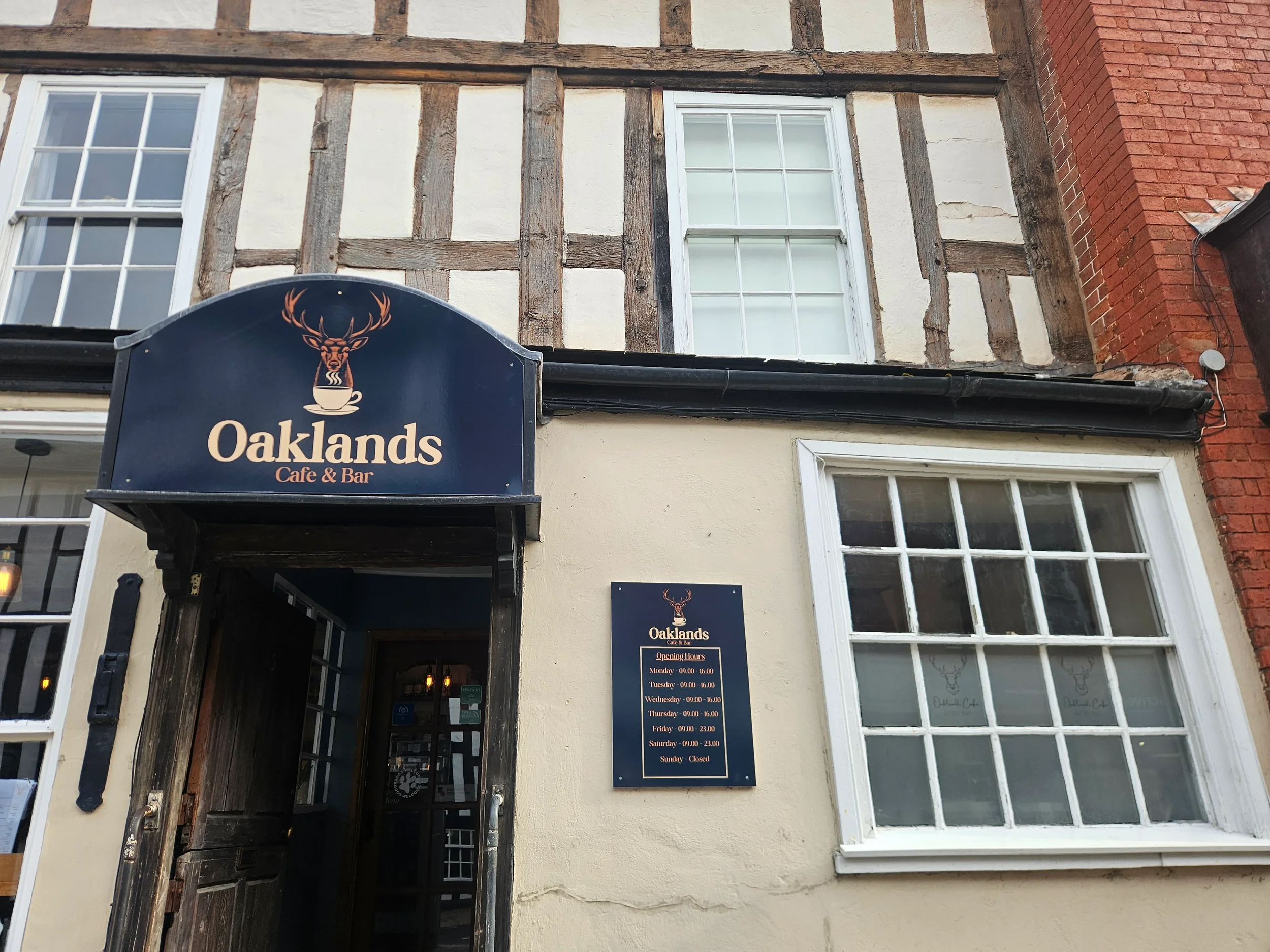 Exterior of a building with a sign for Oaklands Café & Bar, showing decorative half-timbered architecture, two white-framed windows, and a black hanging sign with a deer and coffee cup logo.