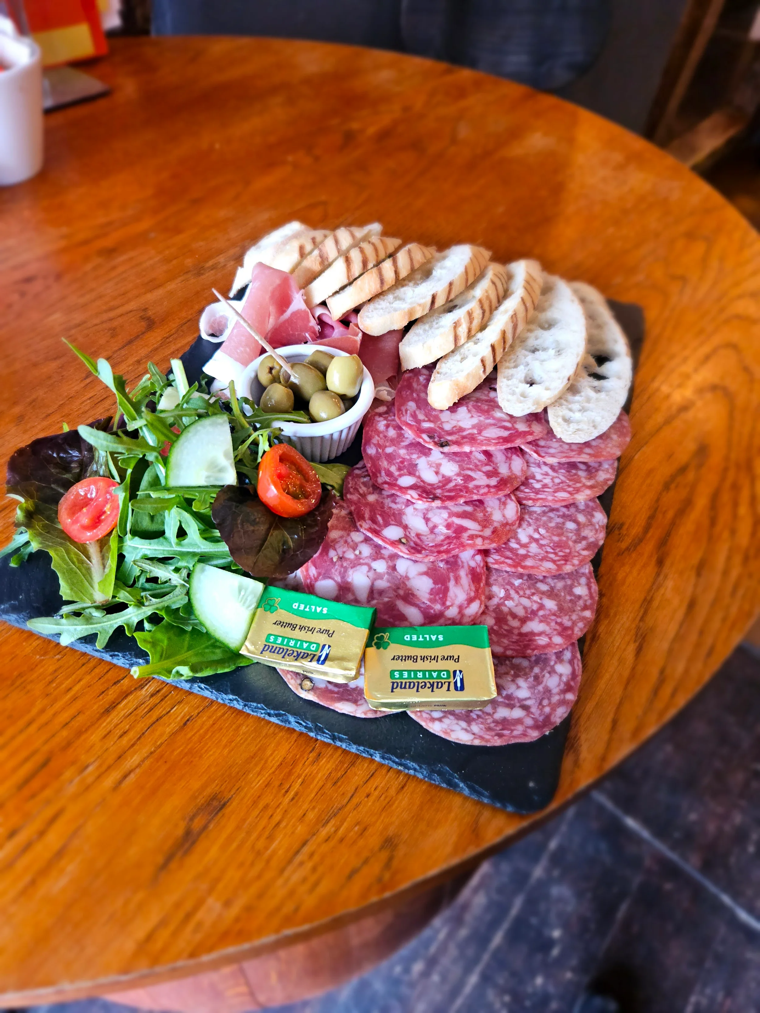 Charcuterie board with sliced meats, cheese, bread, olives, cherry tomatoes, cucumbers, and fresh greens on a wooden table.