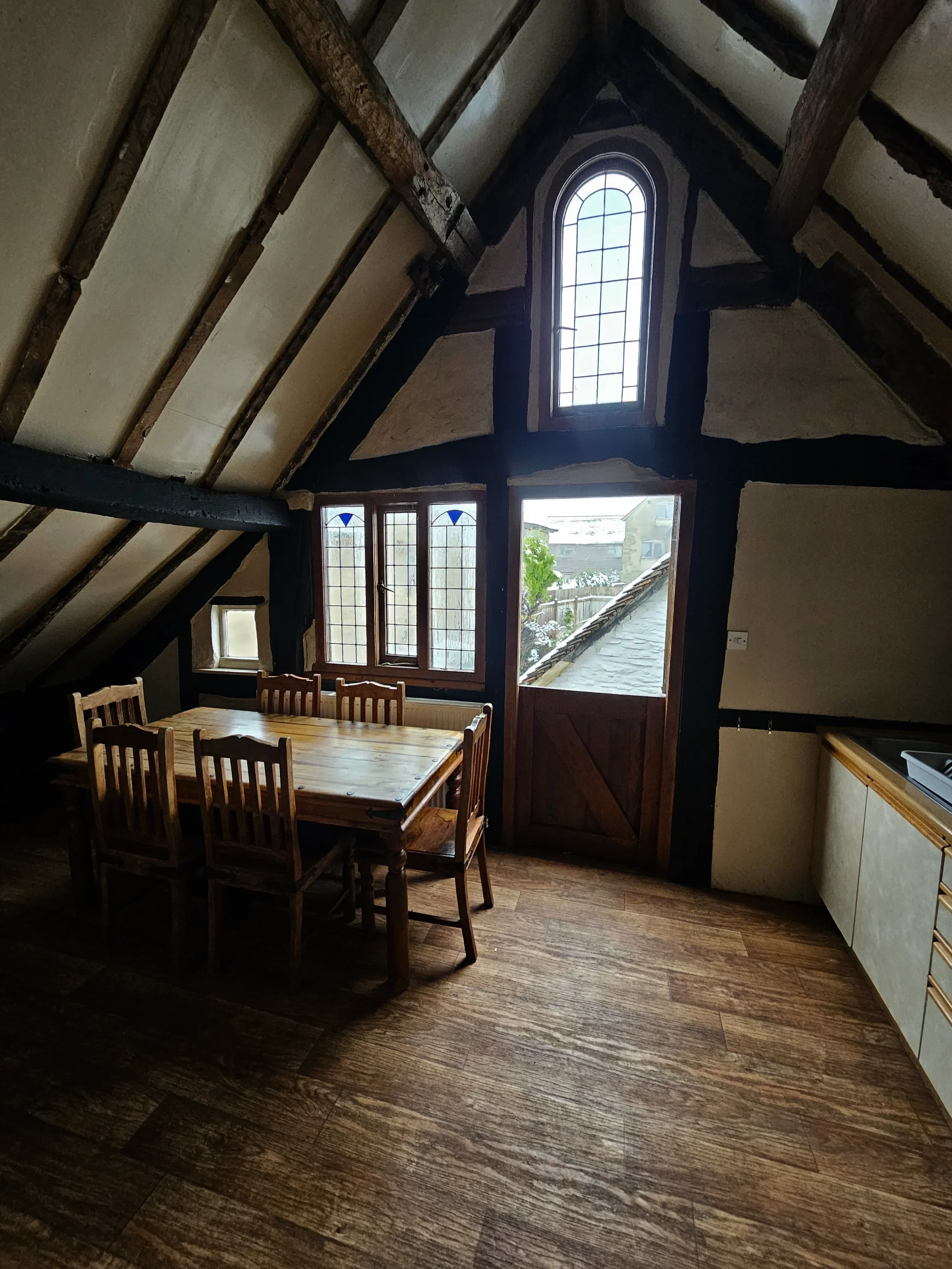 Interior view of a rustic dining area with wooden beam ceiling, a wooden dining table with six chairs, large windows with stained glass, and a view of a garden outside.