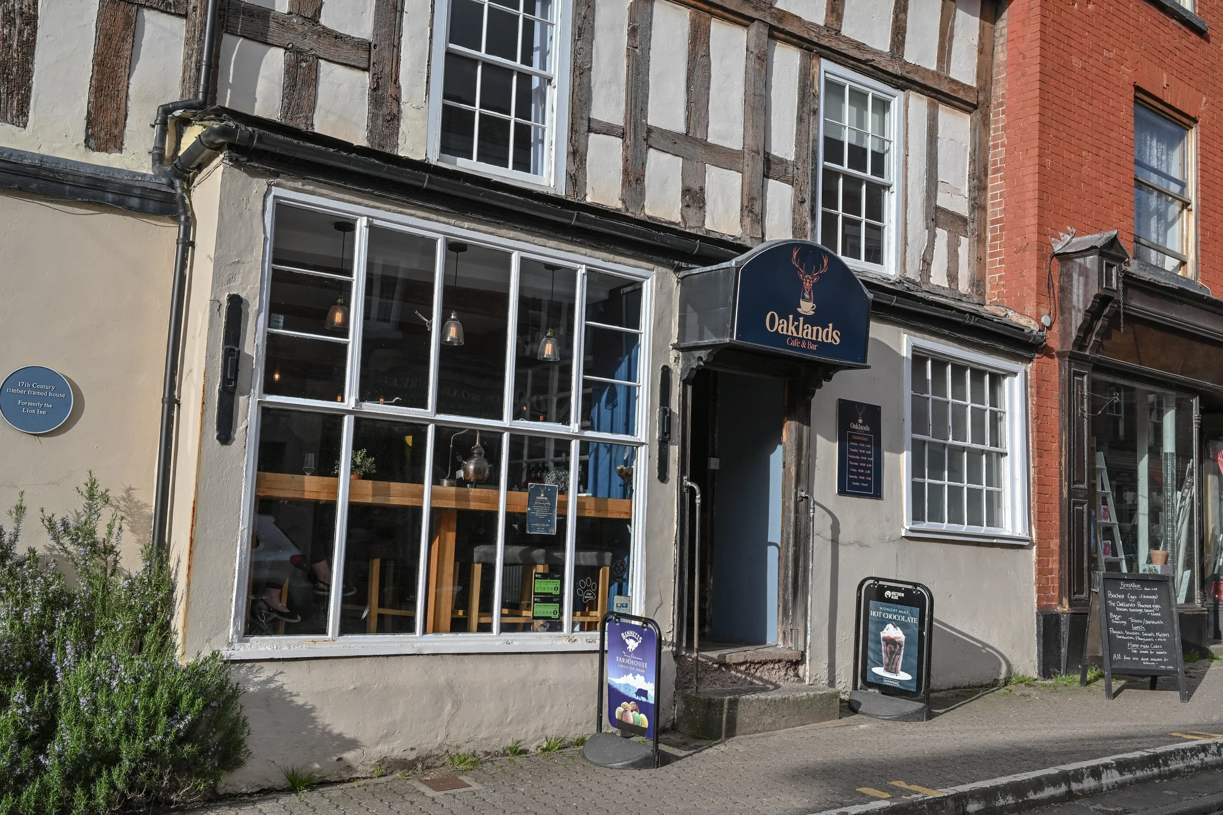 Facade of a historic building with a café and bar named Oaklands, with large windows, a dark blue sign, and outdoor menu boards, on a sunny day.