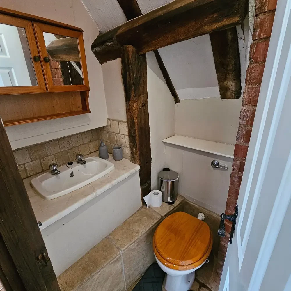 Small bathroom with a white ceramic sink, wooden cabinet, and wooden toilet seat. Exposed wooden beams and brick wall accents.