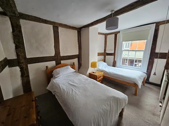 A bedroom with two single beds, a wooden nightstand with a yellow lamp, a window with a white shade, and exposed wooden beams on the ceiling and walls.