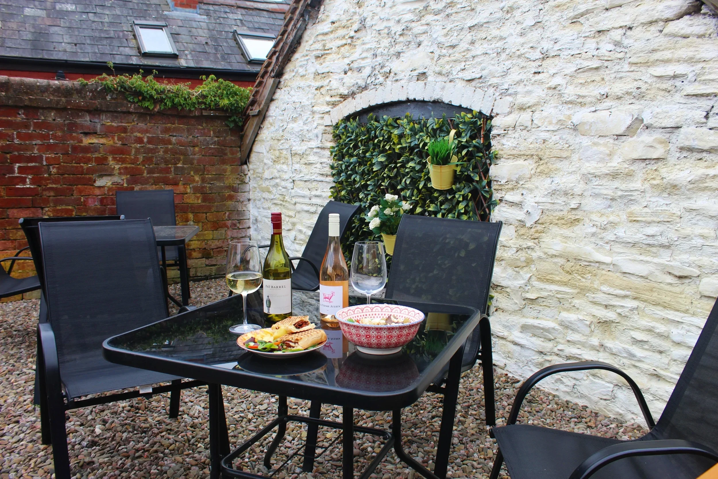 Outdoor dining table set with wine bottles, glasses, a bowl of salad, and bread, surrounded by black chairs, against a background of white and brick walls with potted plants.