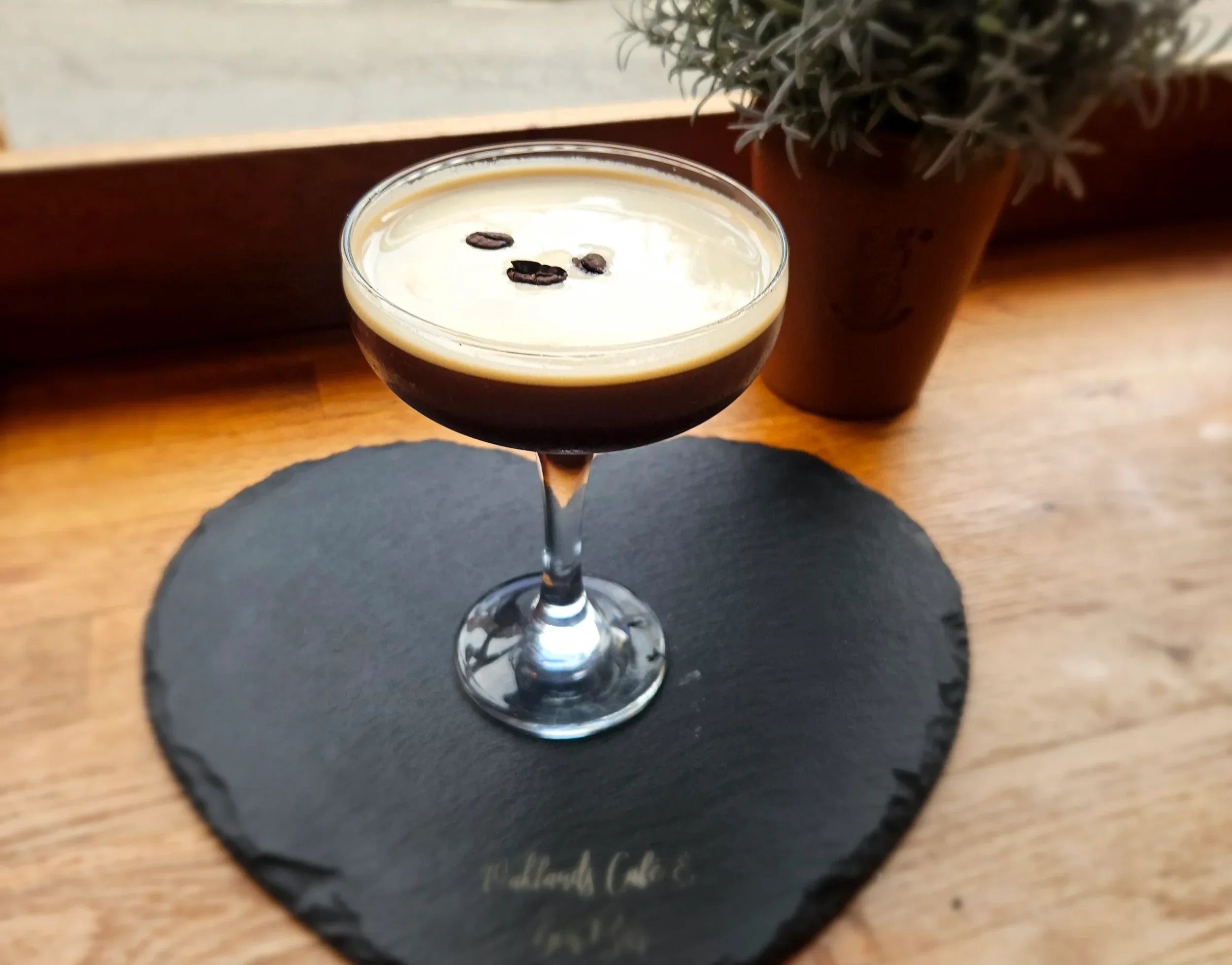 A cocktail glass filled with a dark coffee-based drink, topped with a foam layer and three coffee beans, placed on a round black coaster on a wooden table, with a pot of lavender plant in the background.