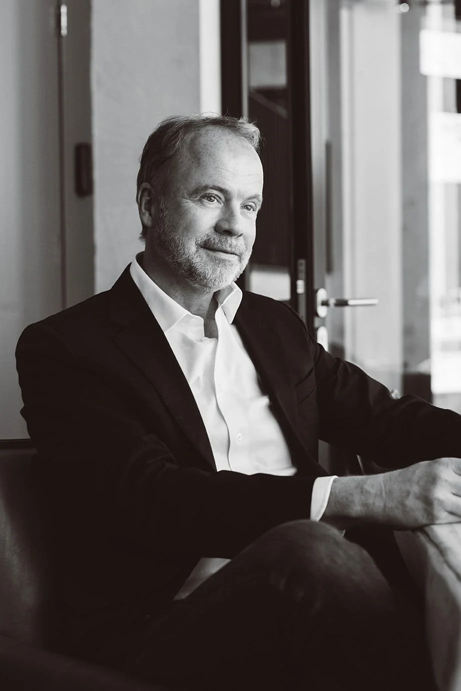 A black-and-white portrait of a middle-aged man with a beard, wearing a suit jacket and white shirt, sitting in an indoor setting.