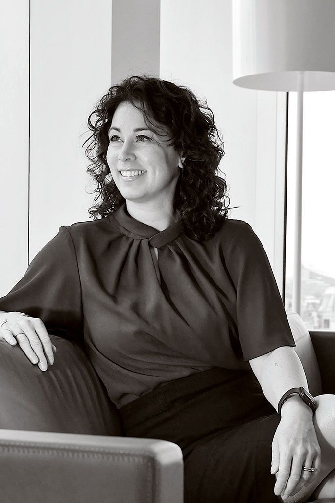 A woman with curly hair sitting on a sofa, smiling, in a bright room with large windows and a floor lamp.
