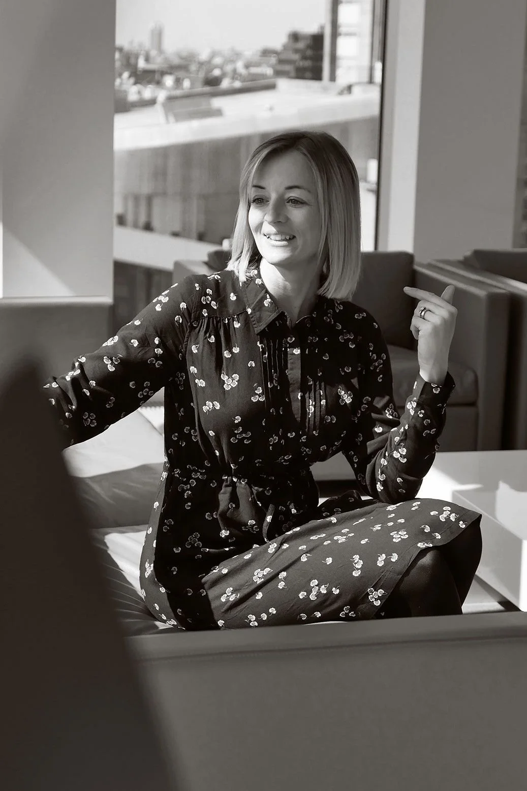 A woman with shoulder-length hair sitting on a bed in a room with a window, wearing a patterned dress, smiling and gesturing with her right hand.