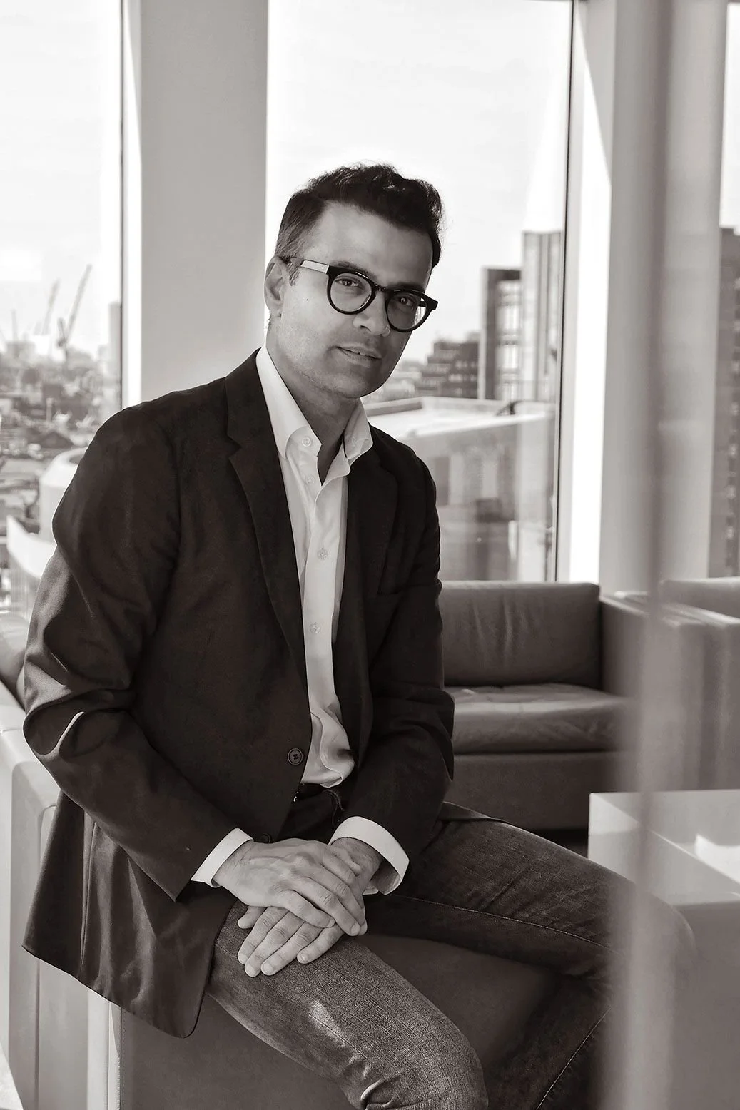 A man in glasses wearing a black blazer and white shirt, sitting in a modern office with large windows in the background.