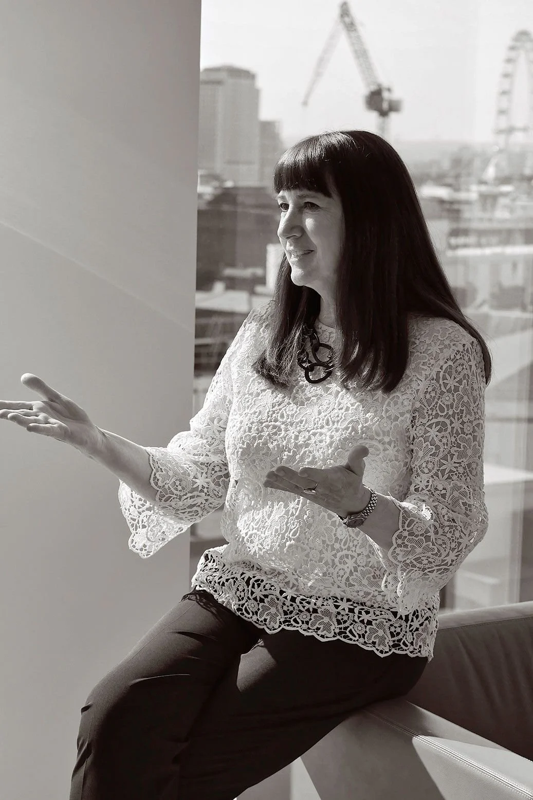A woman with dark hair and bangs, wearing a lace blouse, sitting and gesturing with her hands during a conversation or presentation indoors with city buildings and cranes visible in the background.