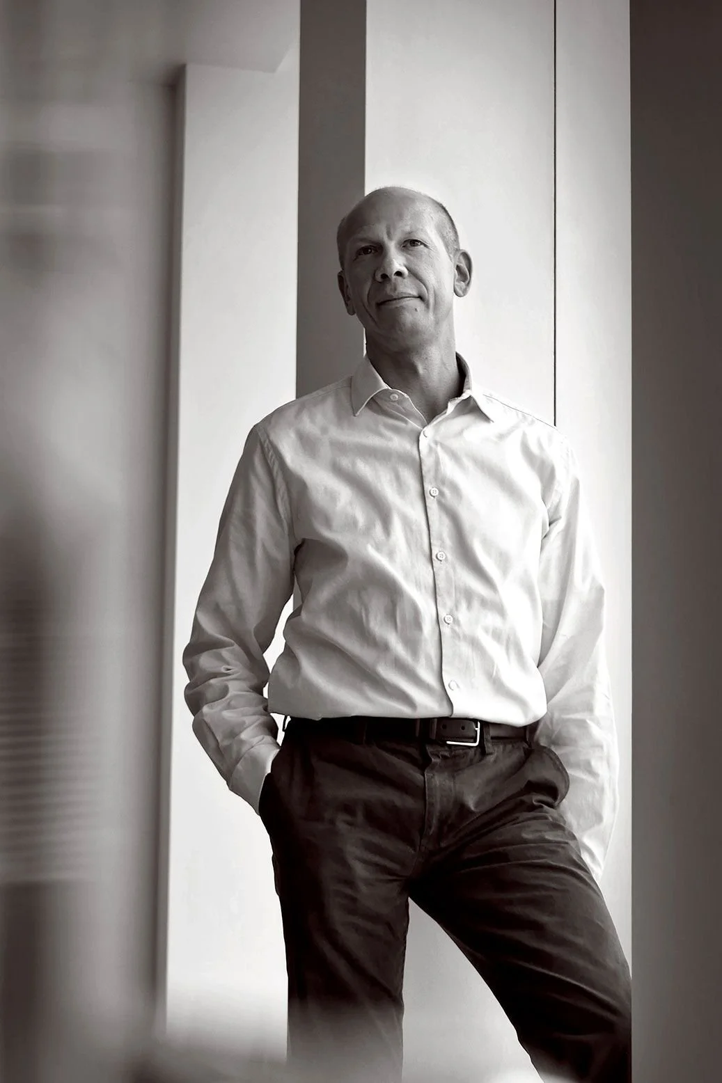 A black-and-white photo of a man in a white dress shirt and dark pants, standing with one hand in his pocket, leaning against a wall in a modern, minimalistic interior.