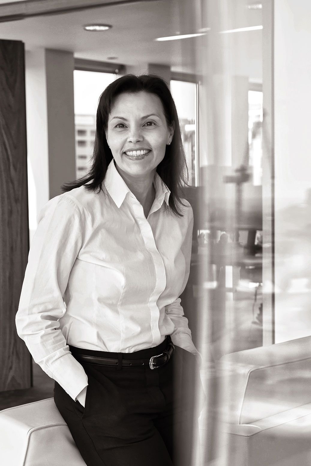 A woman with dark hair smiling, wearing a white button-up shirt and black pants, standing in an office environment near a glass wall.