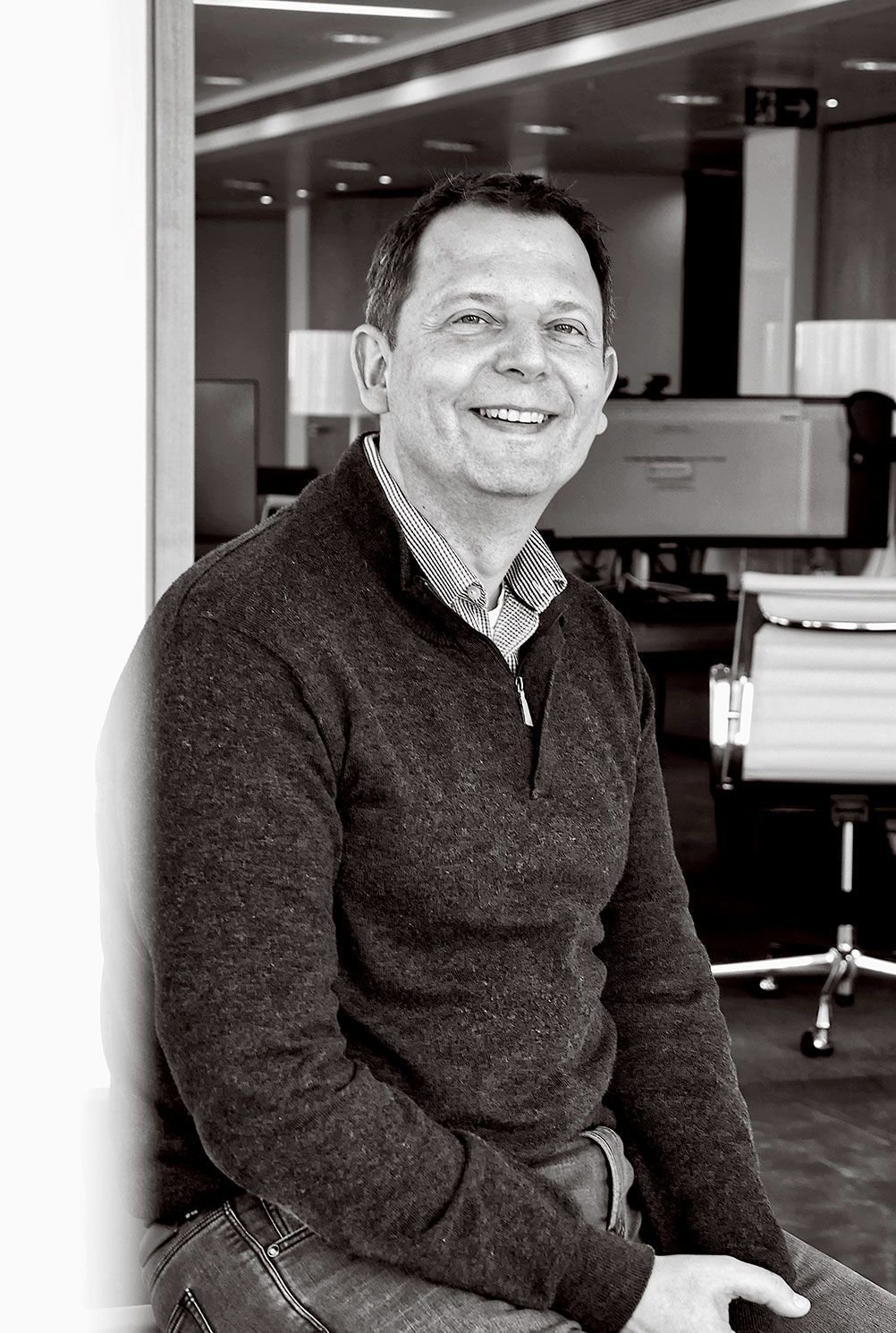 A smiling man sitting in an office, wearing a dark sweater and a collared shirt, with office furniture and computer monitors in the background.