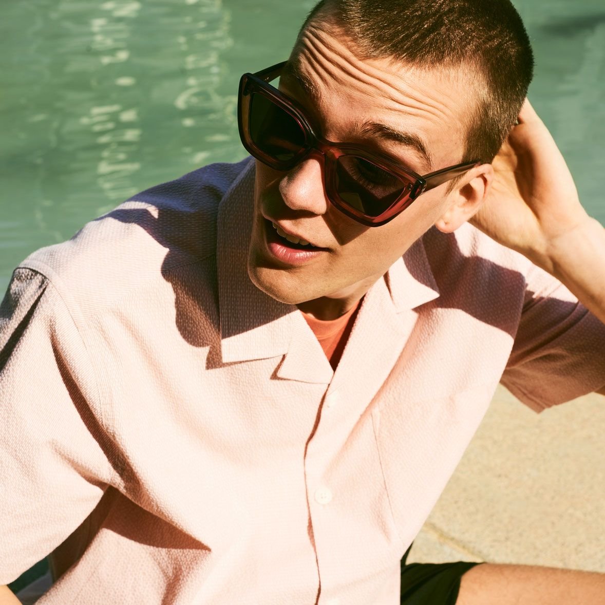 A man wearing large sunglasses, a light pink short-sleeved shirt, and sitting by a body of water, looking to the side with one hand behind his head.