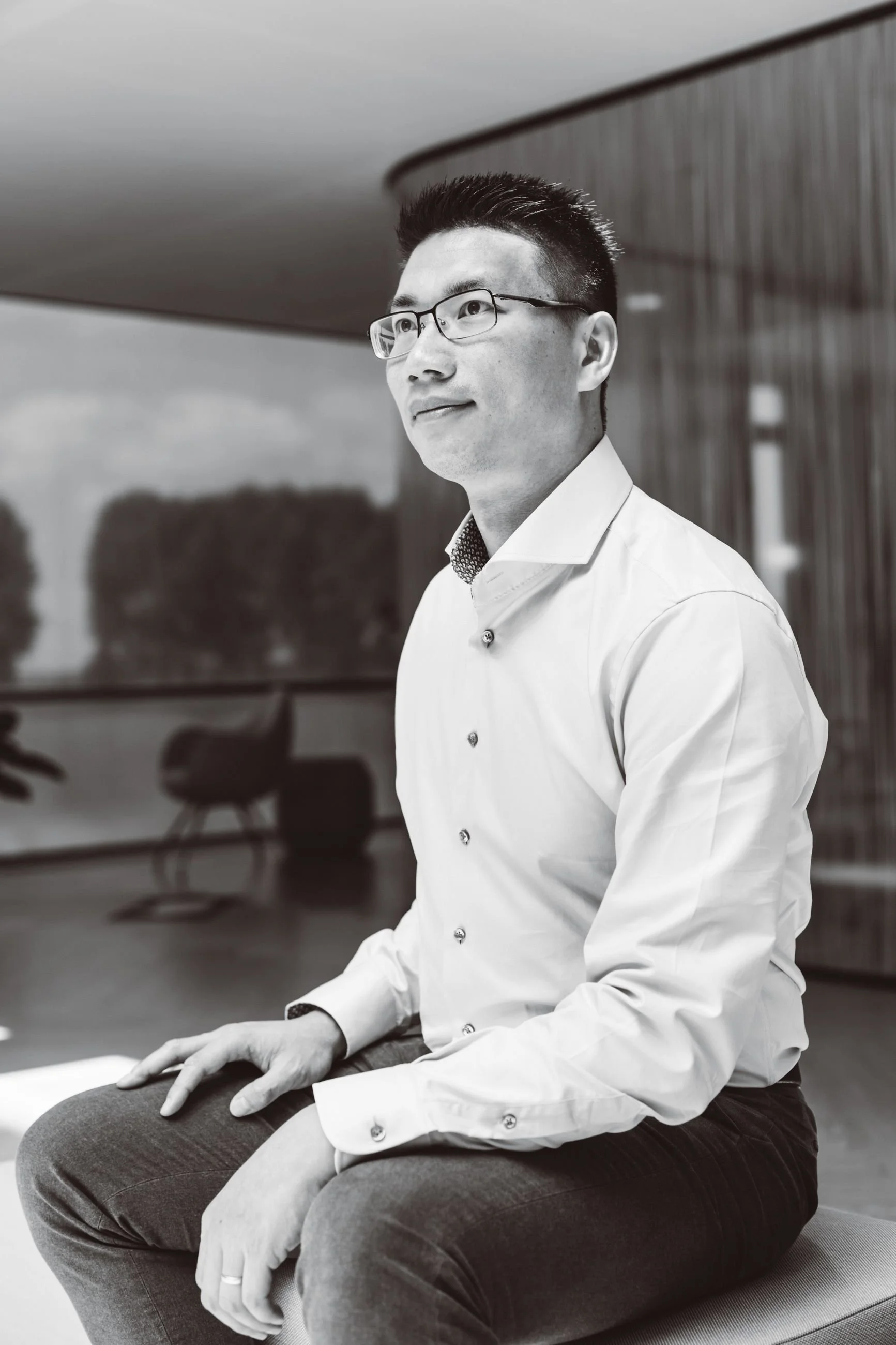 A man with glasses wearing a dress shirt and dark pants, seated on a bench in a modern office or conference room. The background includes wood paneling and blurred chairs.