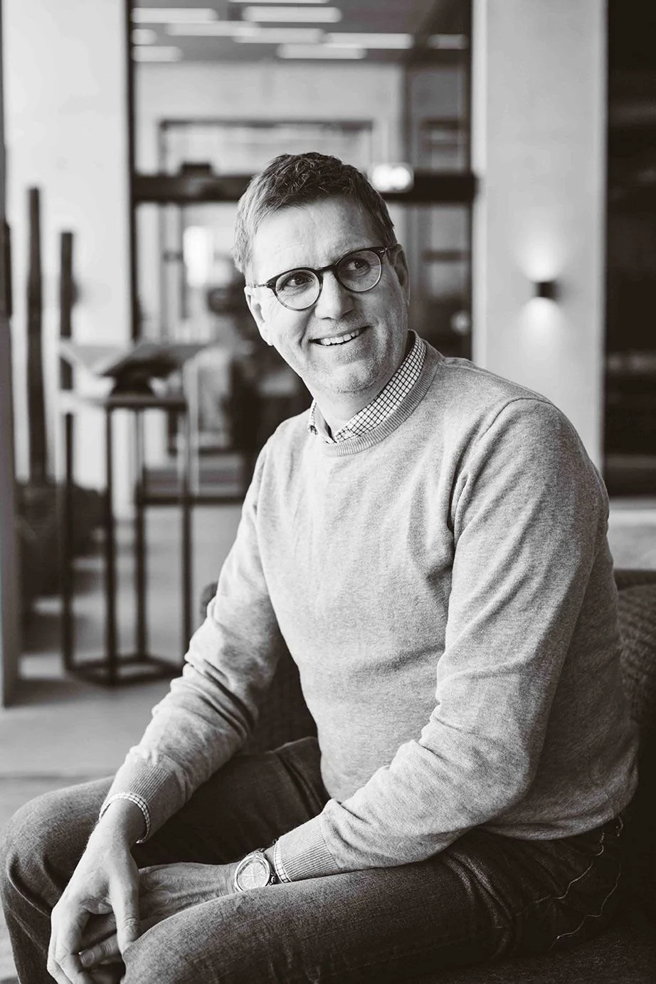 A smiling man with glasses and short hair sitting indoors in a modern, well-lit space.