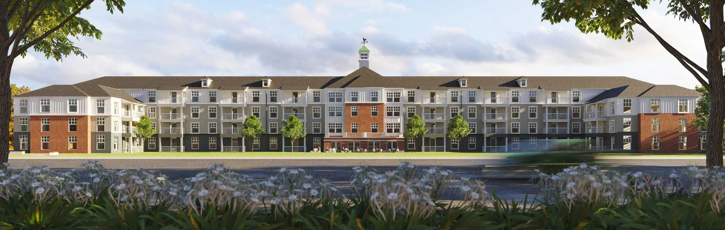 A large multi-story apartment building with a mix of brick and white siding, surrounded by green trees and grass, with a flower bed in the foreground.