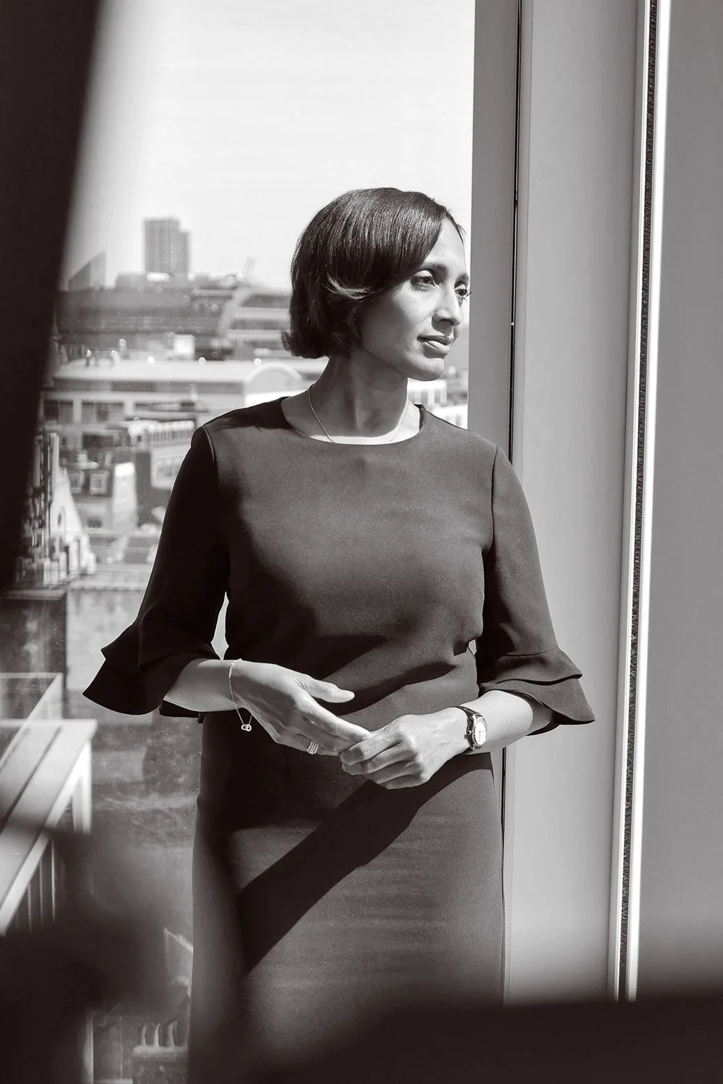 A woman with short, dark hair looking out a window with a cityscape behind her.