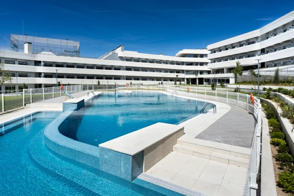 Empty outdoor swimming pool with a modern white building in the background, under a clear blue sky.