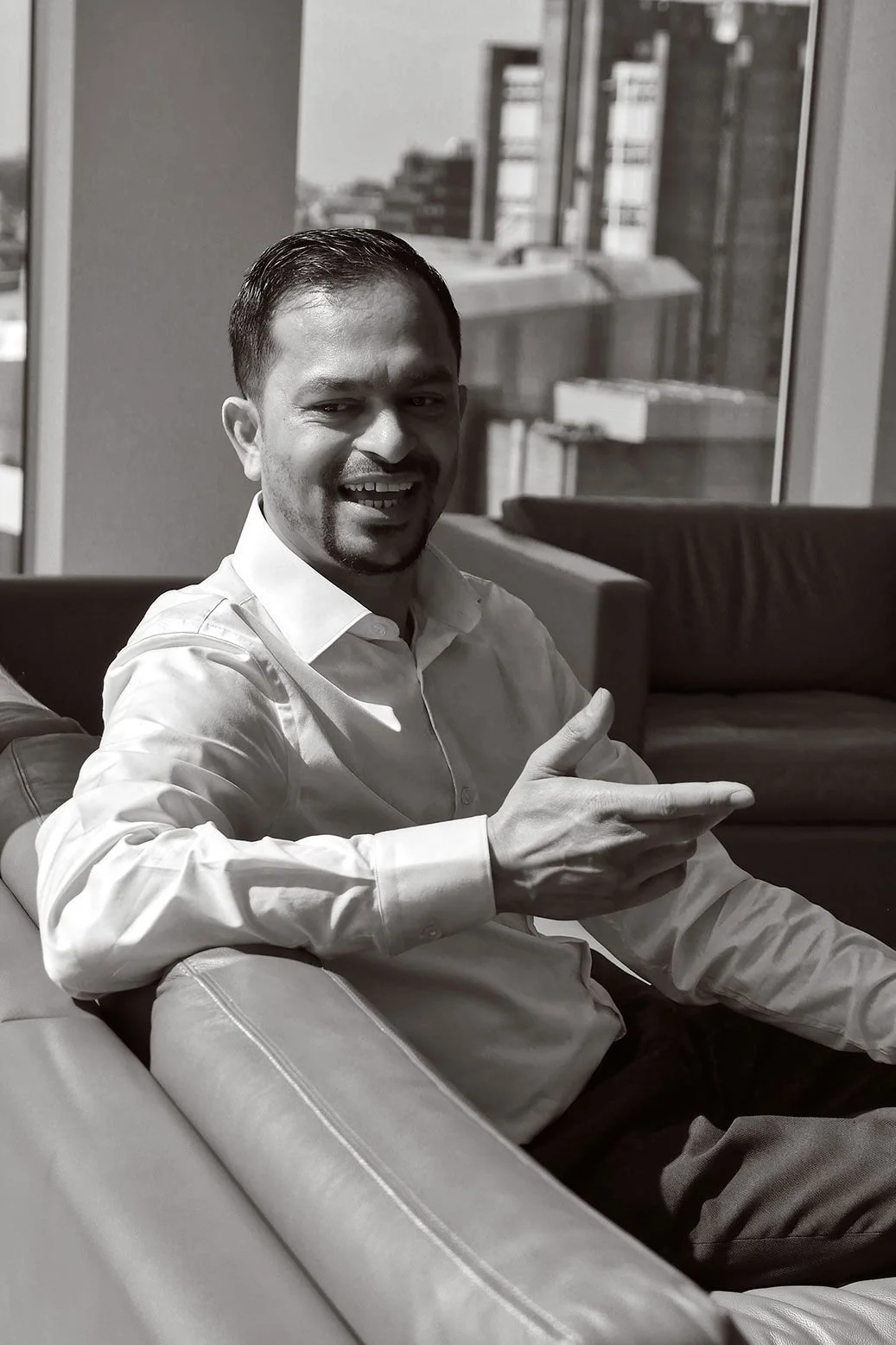 A man in a white dress shirt sitting on a couch, smiling and pointing with his right hand. The background shows large windows with a cityscape view, and a bookshelf.