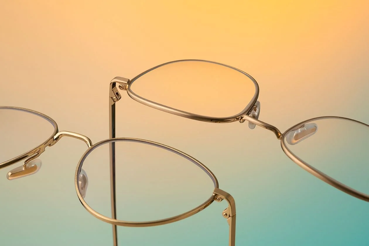 Multiple pairs of round glasses with thin metal frames hanging on a stand against a pastel gradient background.