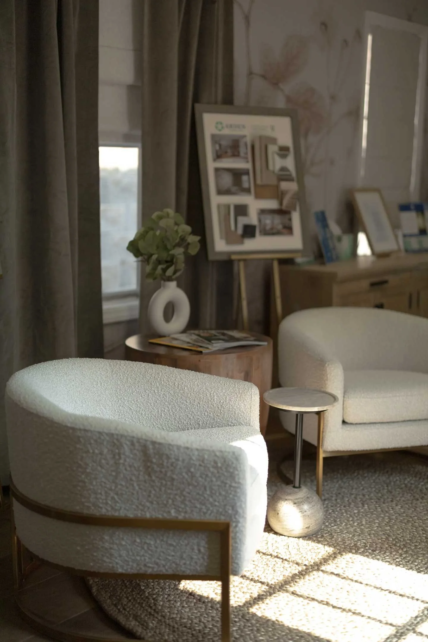 A cozy corner of a room with two white armchairs, a small side table with a textured base, a lamp, a photo frame, a flower arrangement, and a wooden dresser with framed pictures, all lit by natural sunlight.