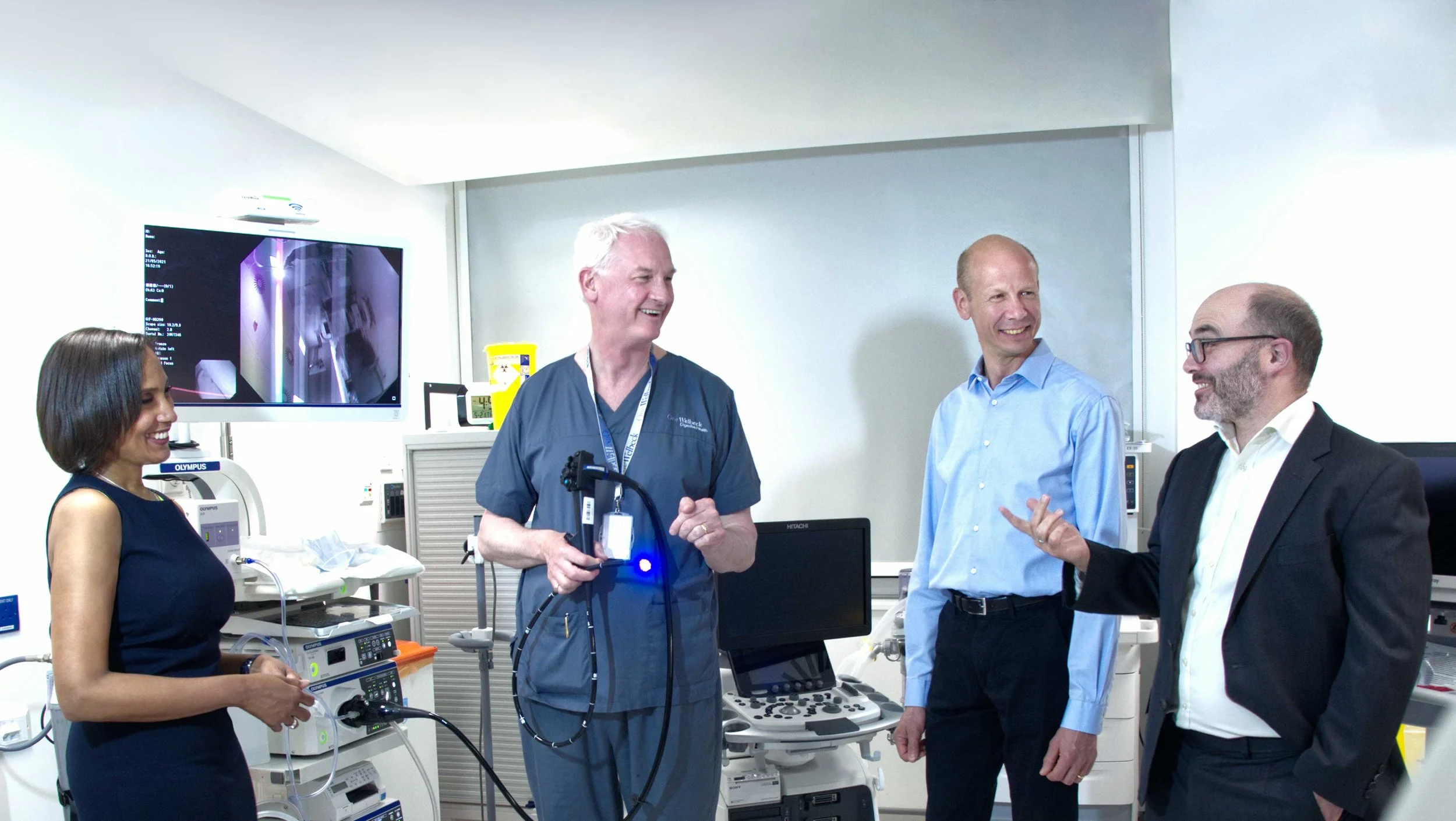 Group of four professionals, three men and one woman, engaged in a conversation in a hospital or medical setting with medical equipment around, including monitors and medical devices.