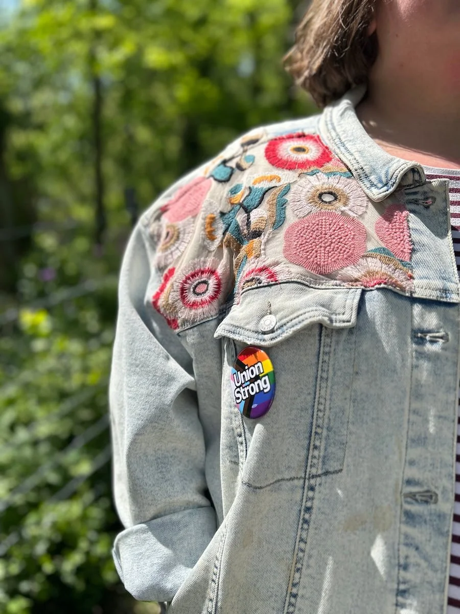 Close-up of a person wearing a light denim jacket with floral embroidery on the shoulder. The jacket has a rainbow-colored rainbow LGBTQ+ pin that reads 'Union Strong.' The background shows greenery.