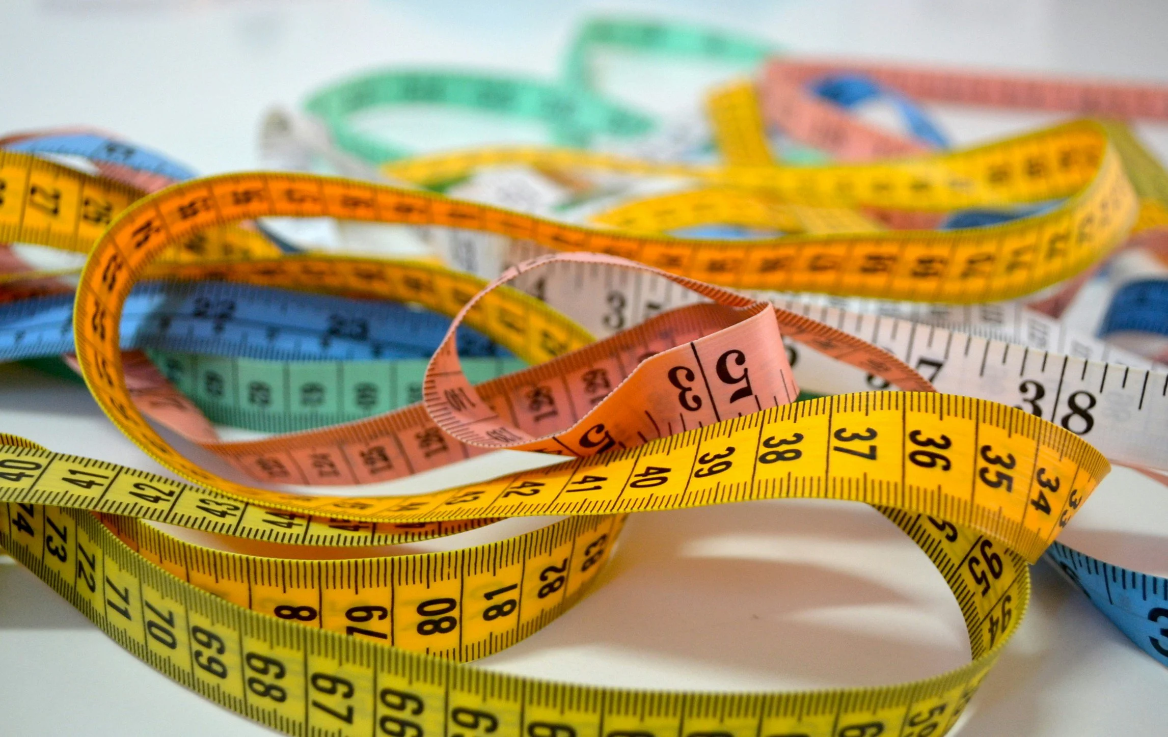 Colorful measuring tapes in yellow, pink, green, blue, and orange tangled on a white surface.