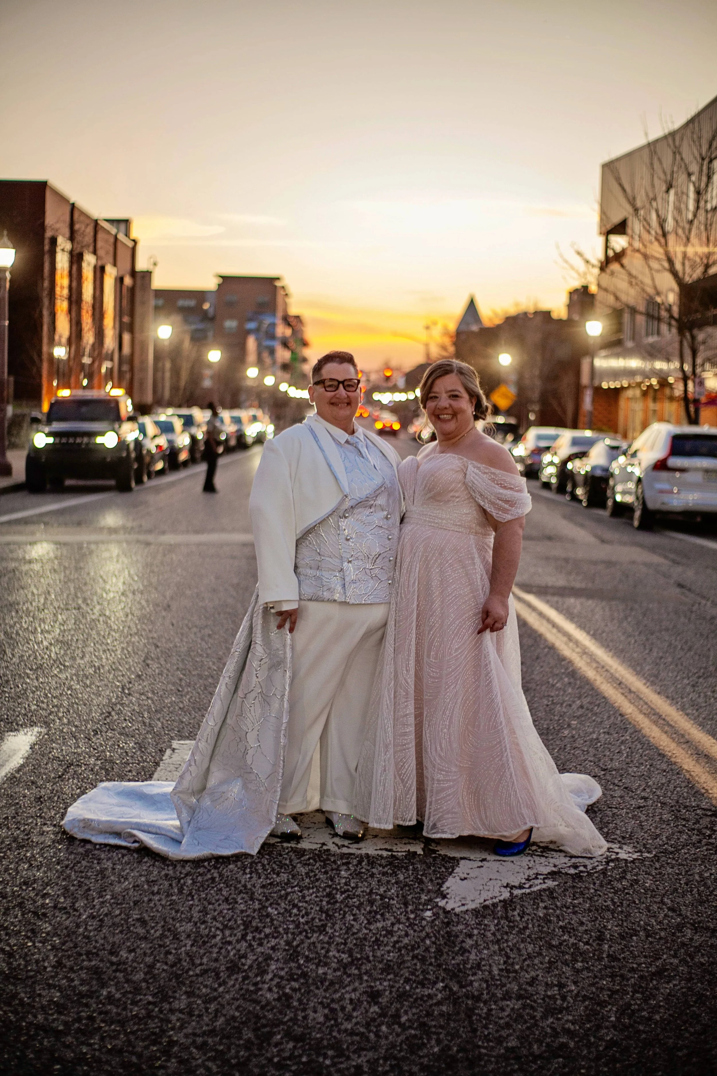 A trans nonbinary person in a custom gender-neutral suit with a train, along with their spouse in a wedding dress, standing in the middle of a street at sunset, with parked cars, buildings, and streetlights in the background.