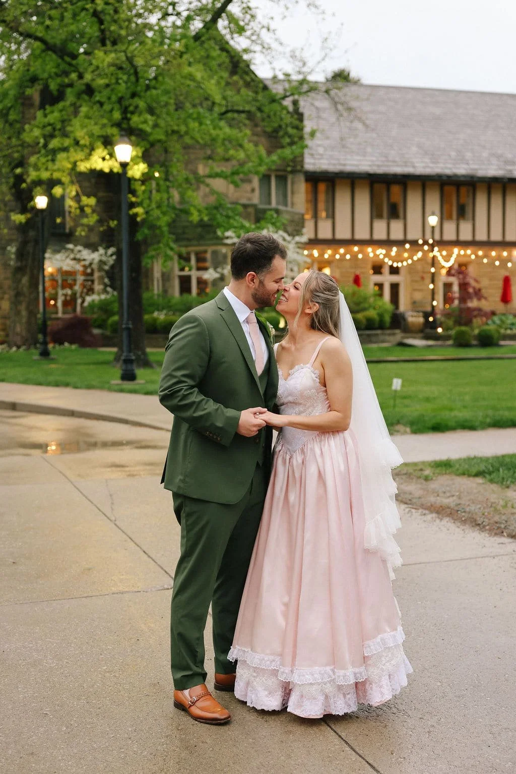 A bridge and groom holding hands and smiling at each other in an outdoor setting with a historic building and string lights in the background, during dusk or evening.