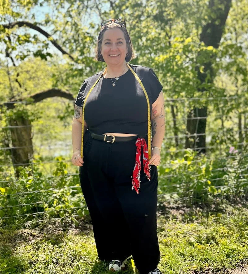 Woman standing outdoors in front of green trees, smiling, wearing a black top and pants, a yellow measuring tape around her neck, sunglasses on her head, a red bandana tied to her belt, and holding her hands in her pockets.