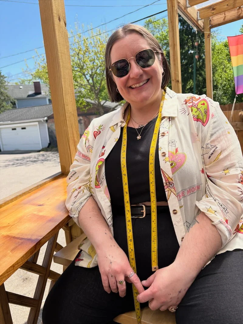 A smiling Molly Walz with short brown hair wearing sunglasses, a black shirt, and a patterned white shirt with colorful designs. She has a measuring tape around her neck sitting outdoors on a wooden patio with a pride flag in the background.