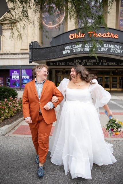 Two joyful people walk arm-in-arm outside the Ohio Theatre, with a marquee displaying 'Welcome Solo,' 'Gameday,' and 'Love Story.' One person wears a white wedding dress holding a bouquet, while the other wears an orange suit and gray shoes.