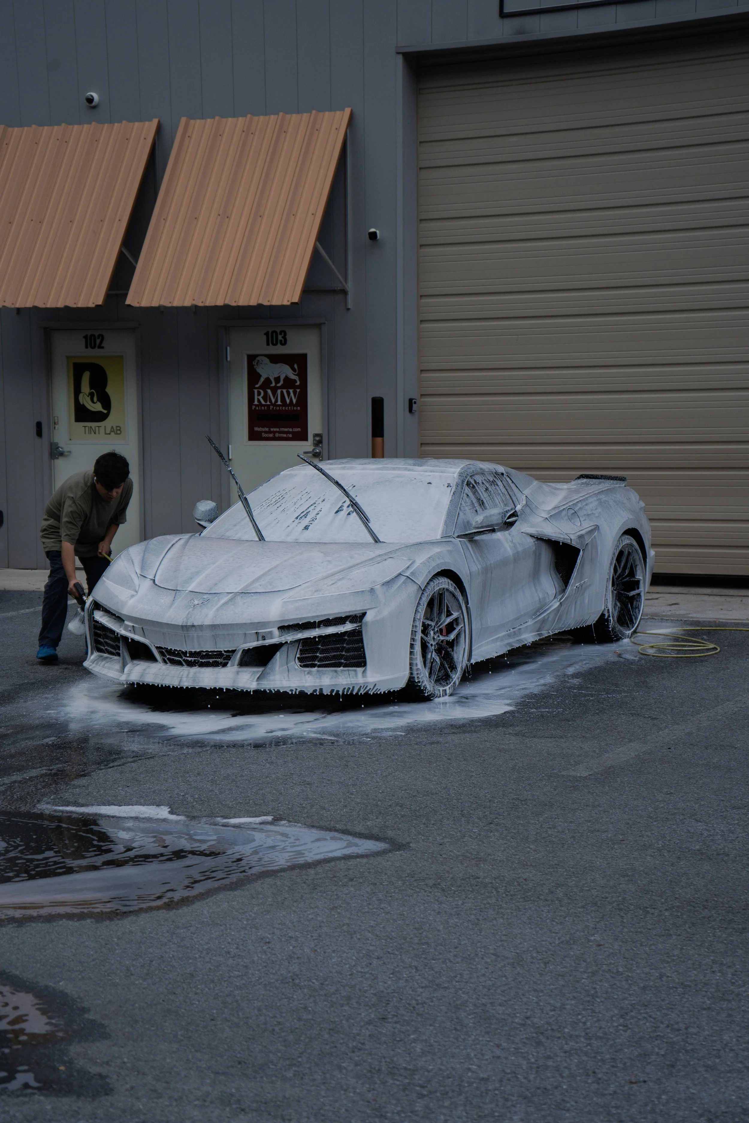 A man washing a white sports car covered in soap suds outside a building with gray walls, two orange awnings, and a large garage door.