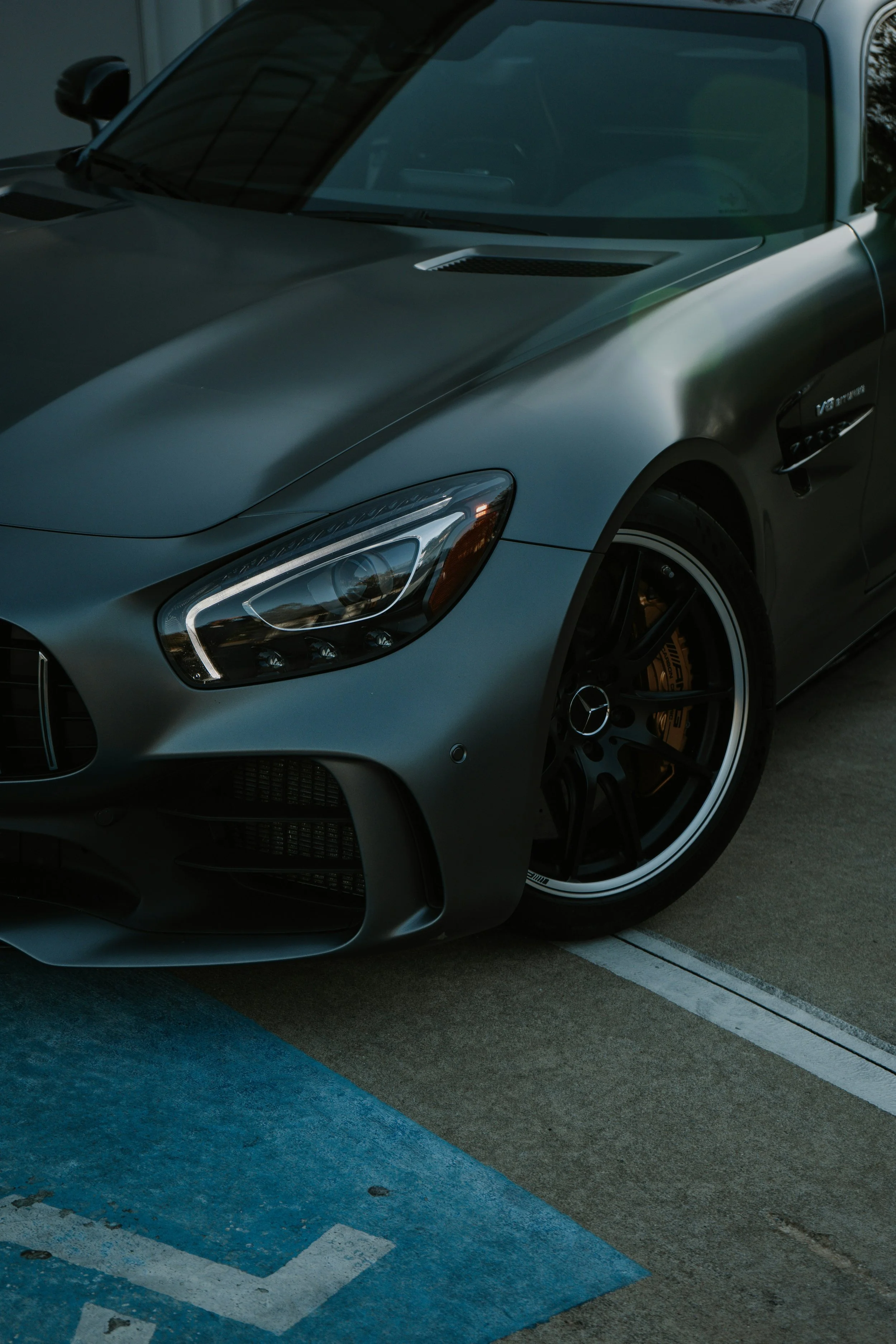 A silver Mercedes-Benz AMG car parked in a parking space with blue markings, focusing on the front left section of the vehicle showing the headlight, wheel, and part of the hood.