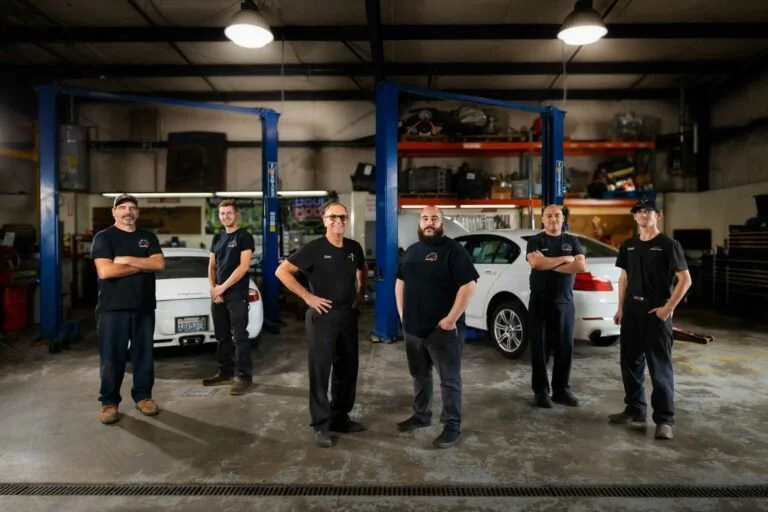 Five mechanics standing in an auto repair shop with lifted cars in the background.