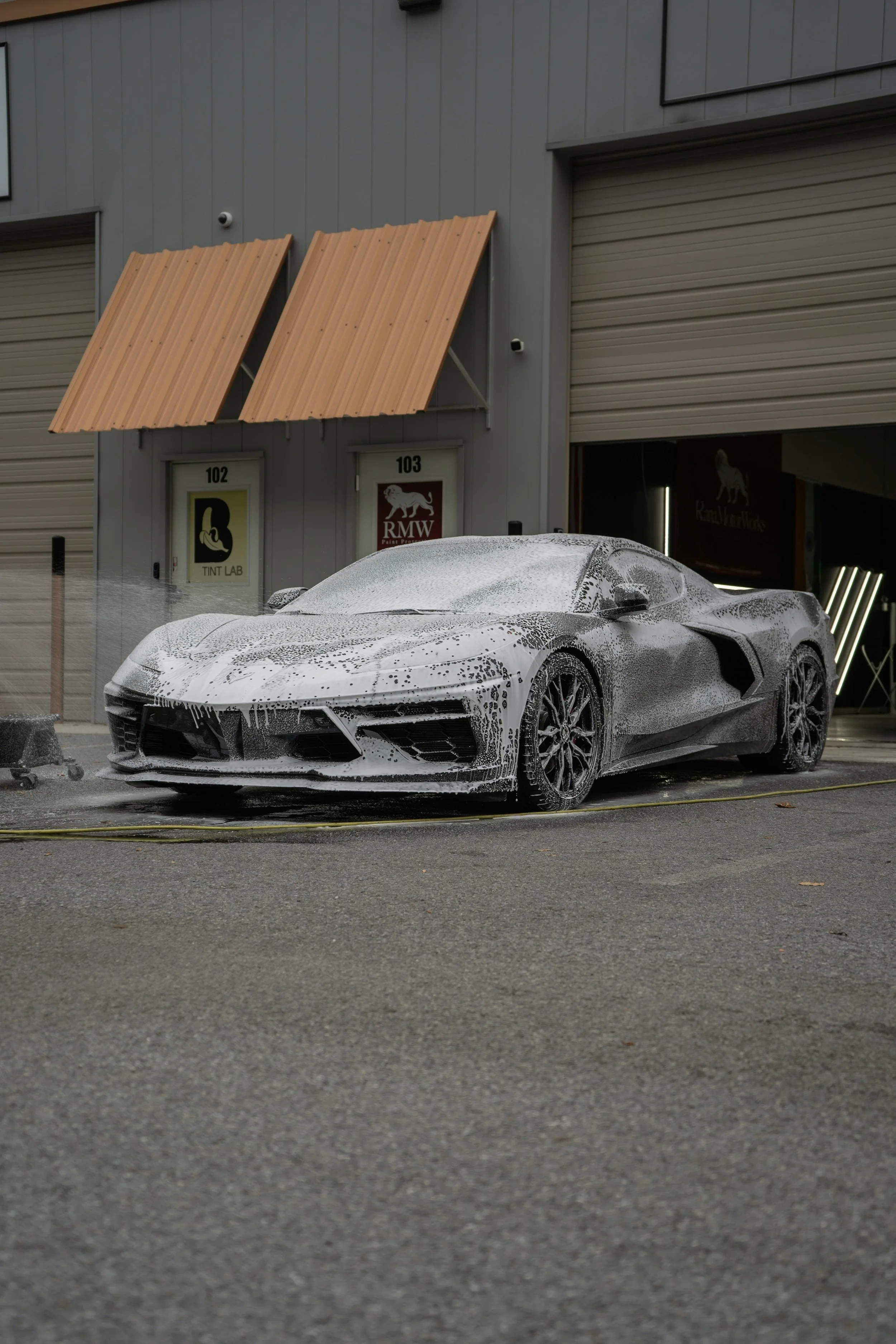 A gray sports car covered in white soap foam, parked outside a commercial building with multiple garage doors and two signs labeled '102 TINT LAB' and '103 RMW'.