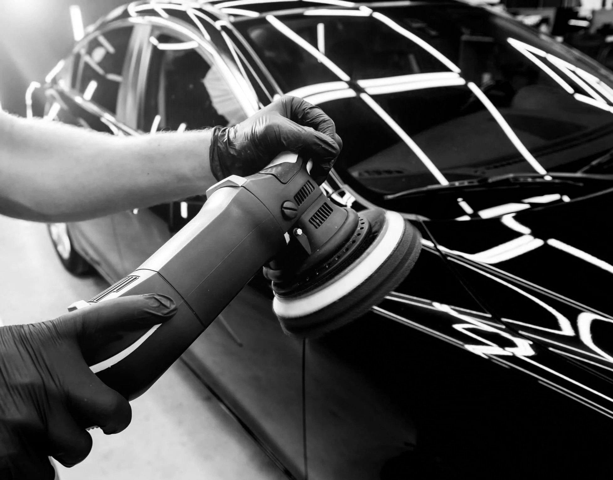 A person wearing gloves polishes or buffs a black car with a handheld polisher in a garage or workshop.