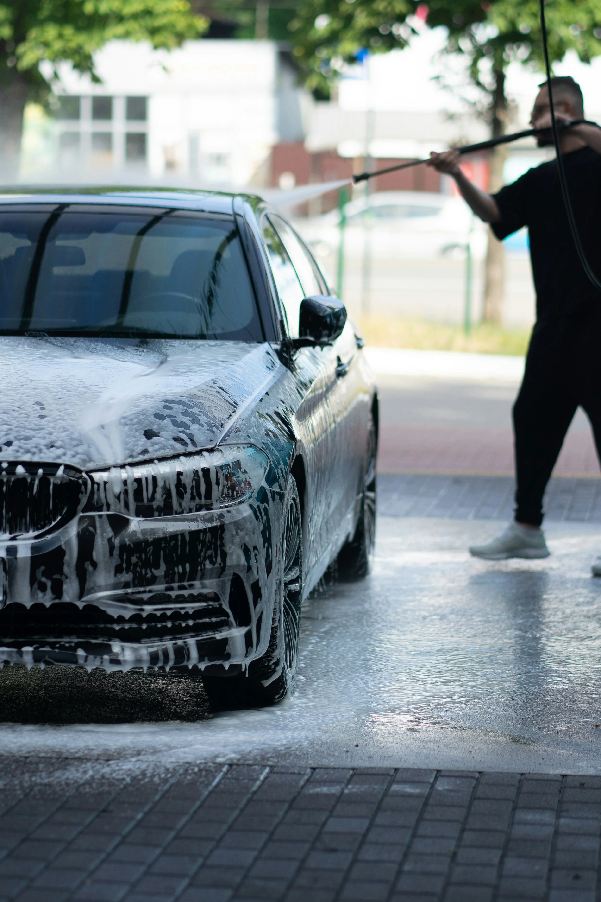 A person is washing a black car with a hose, creating soap bubbles on the vehicle. The scene takes place outdoors with some trees and a building in the background.