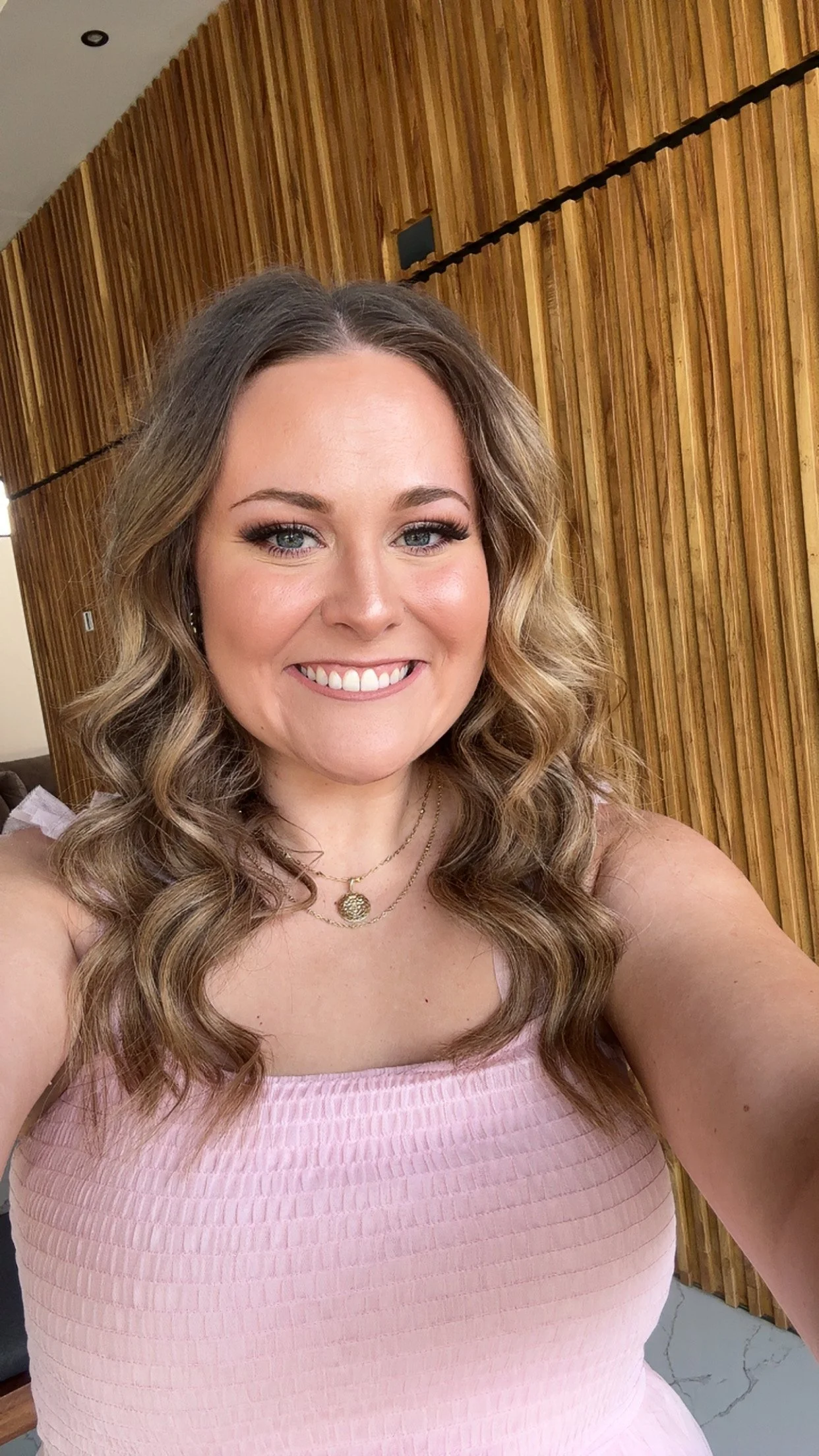 A smiling woman with wavy blonde hair wearing a pink dress and layered necklaces, taking a selfie indoors with wooden paneling in the background.