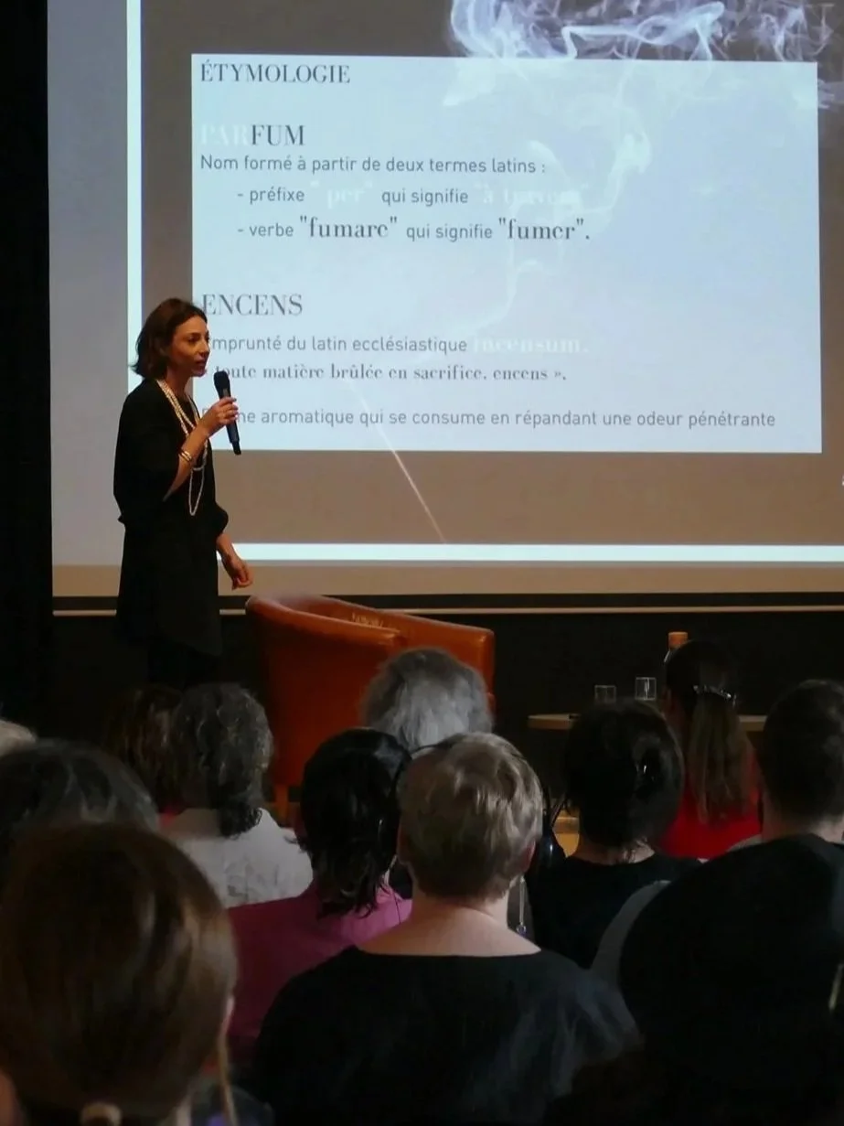 A woman giving a presentation on the etymology of the word "perfume" to an audience, with a slide displaying text in French on a large screen behind her.