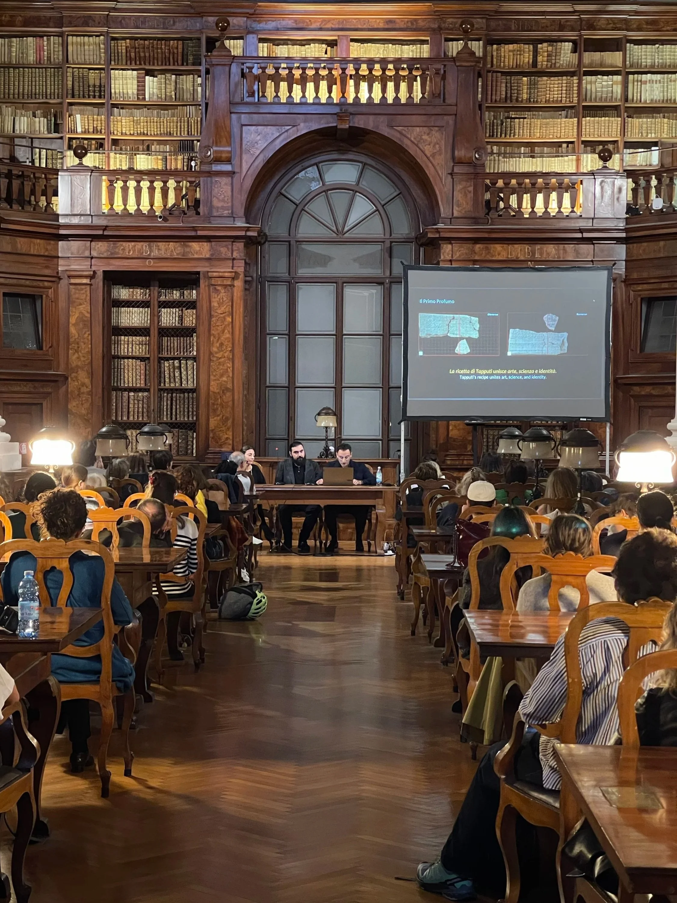 A large, ornate library room with wooden bookshelves filled with old books, a high ceiling, and a group of people seated in chairs, attending a presentation or lecture. There is a large screen displaying a presentation in front of the room.