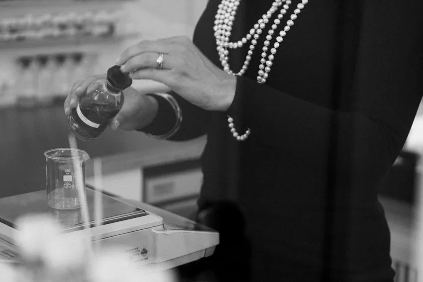 Person pouring liquid from a bottle into a beaker on a digital scale, wearing a black shirt and pearl necklaces, in a laboratory setting.