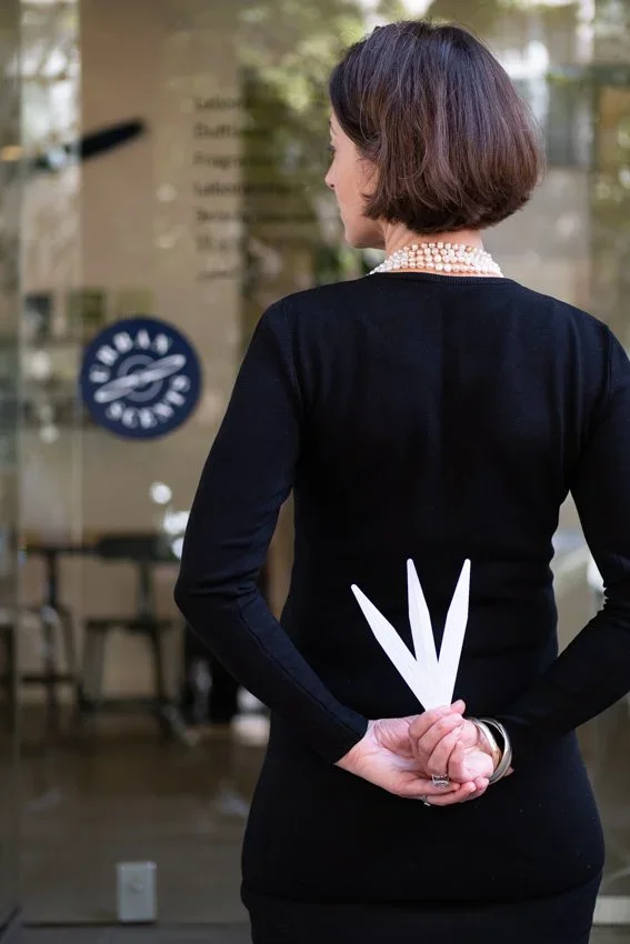 A woman with short brown hair wearing a black dress and pearl necklace, standing with her back to the camera inside a building, holding perfume testing strips behind her back.
