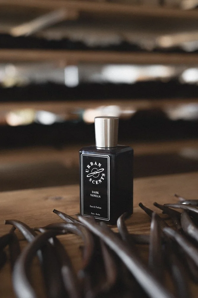 A blue bottle of Urban Scents Dark Vanilla perfume on a wooden surface, surrounded by natural vanilla beans.