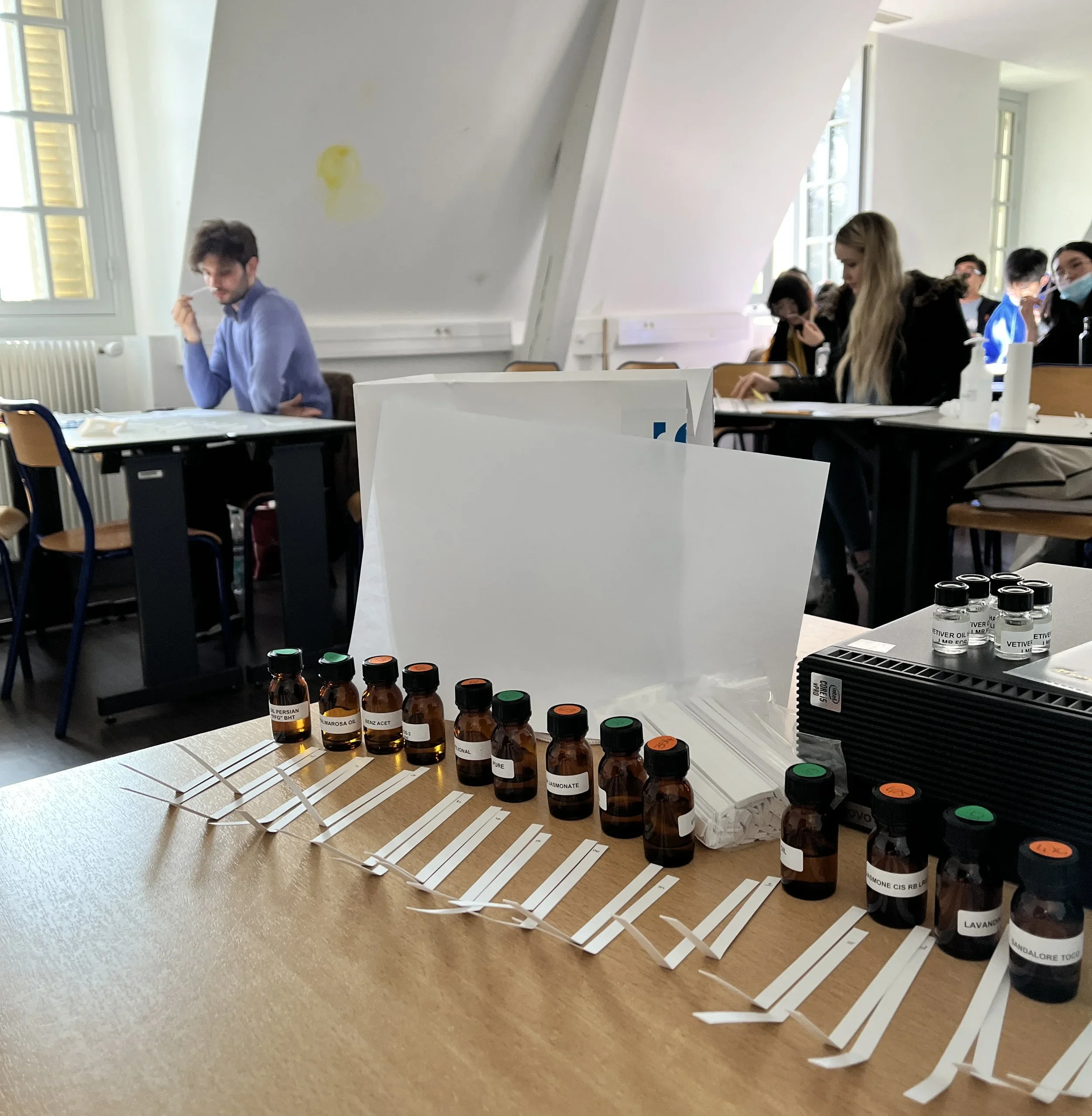 A table with samples of essential oils in small bottles, each labeled with different names, and white paper strips with color-coded dots, for testing or identification purposes, in a room with bright natural lighting.